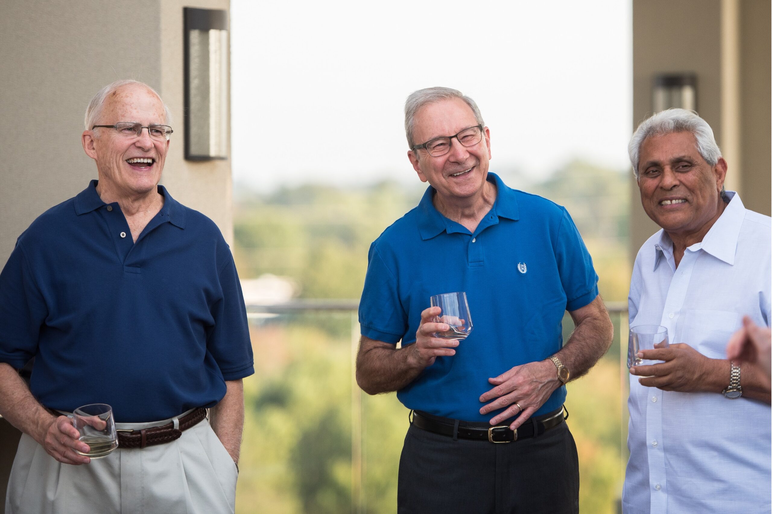Three senior men are laughing while holding drinks on a retirement community balcony.